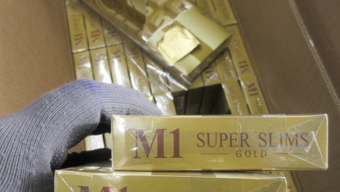 Priekšplānā rokā ar cimdu, paņemtas divas M1 Super Slims Gold cigarešu paciņas, fonā kaste ar cigarešu blokiem