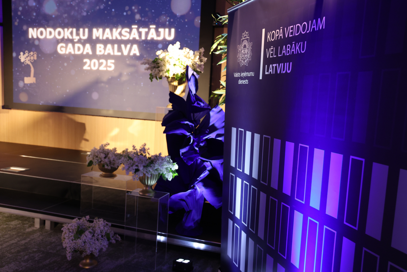 Banneris ar uzrakstu "Nodokļu maksāt'ju gada balva" ziedu dekorācijas
