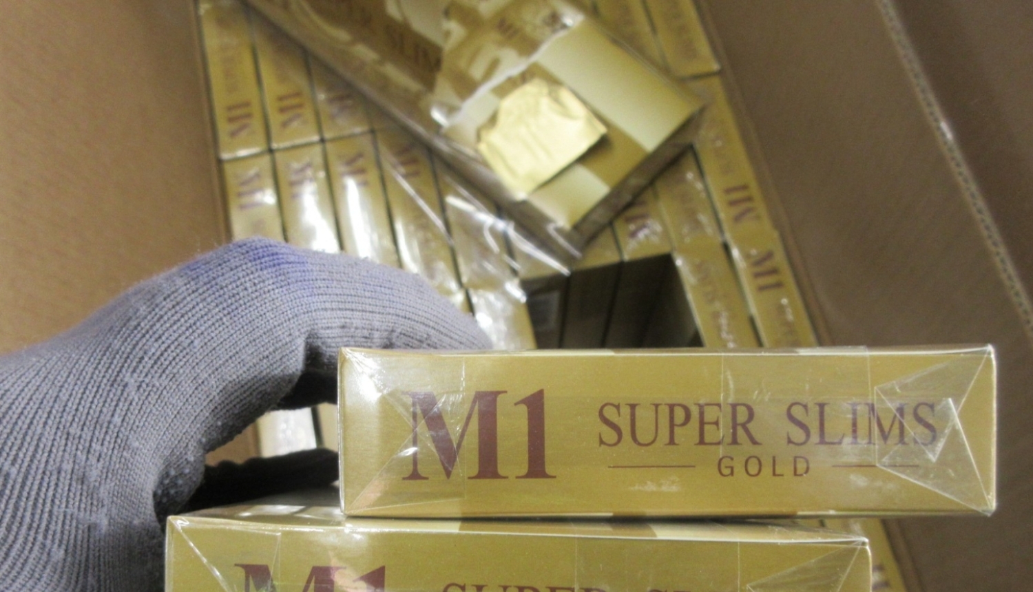 Priekšplānā rokā ar cimdu, paņemtas divas M1 Super Slims Gold cigarešu paciņas, fonā kaste ar cigarešu blokiem