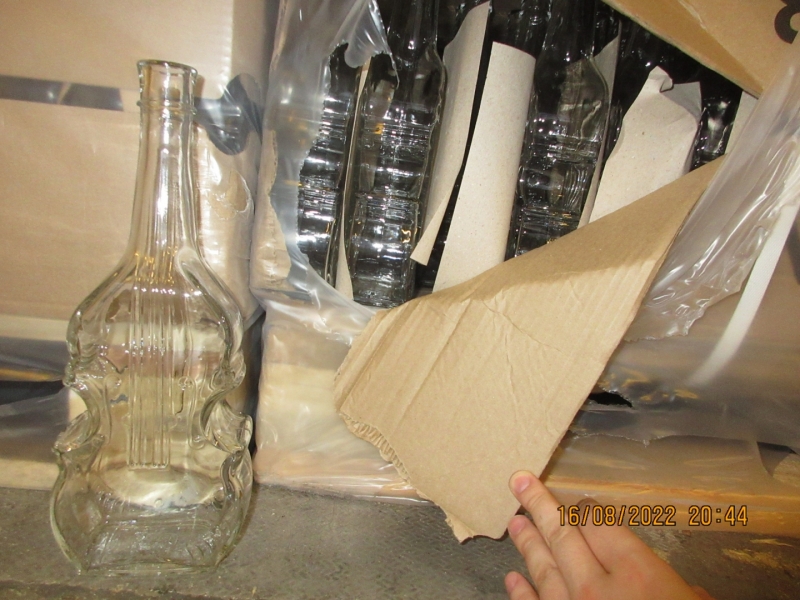 Stikla pudele vijoles formā un fonā kartona kastes ar stikla pudelēm vijoles formā