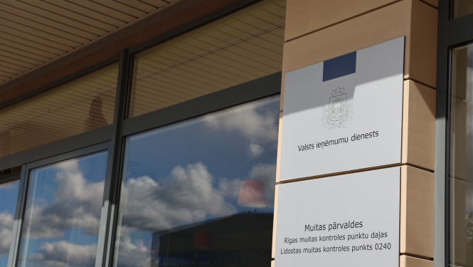 Pie ēkas fasādes piestiprinātas flīzes ar uzrakstiem, logos mākoņu atspīdums