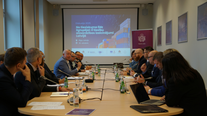 Abās galda pusēs sēž cilvēki, galā ekrāns un banneris