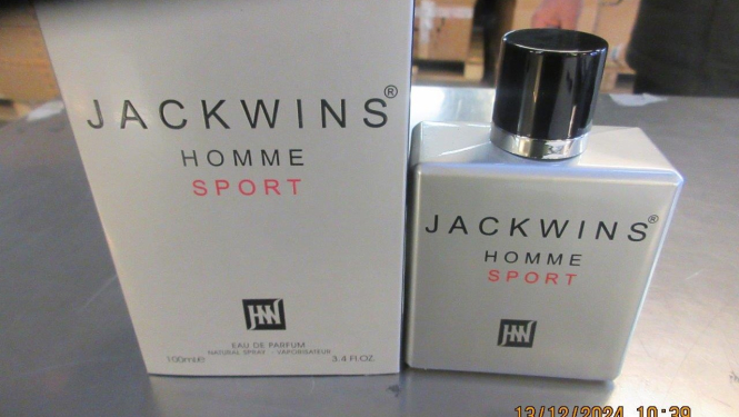 Uz galda pelēka smaržu pudelīte, blakus pelēkam smaržu iepakojumam ar melniem un sarkaniem uzrakstiem 