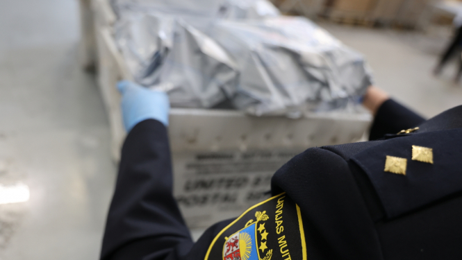 Priekšplānā muitas darbinieka žakete, rokās kaste ar pasta sūtījumiem