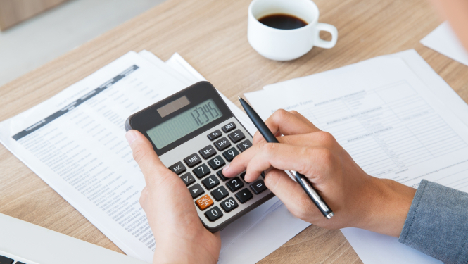 Galda virsma ar papīriem, kafijas krūzi un rokās paņemtu kalkulatoru un pildspalvu 