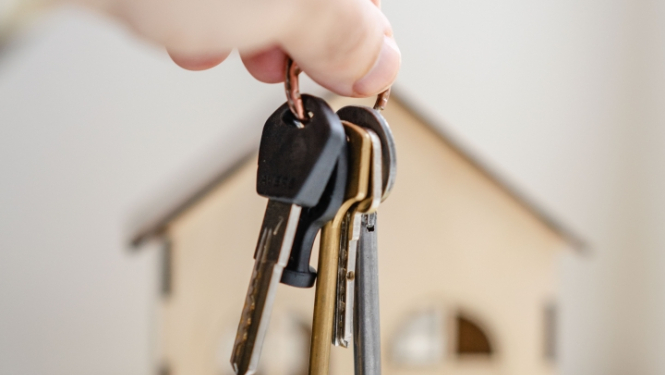 Rokā paņemts atslēgu saišķis, fonā mājas makets