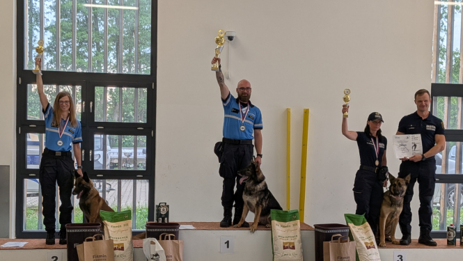 Uz pjedestāla stāv sacensību uzvarētāji – kinologi ar saviem darba suņiem