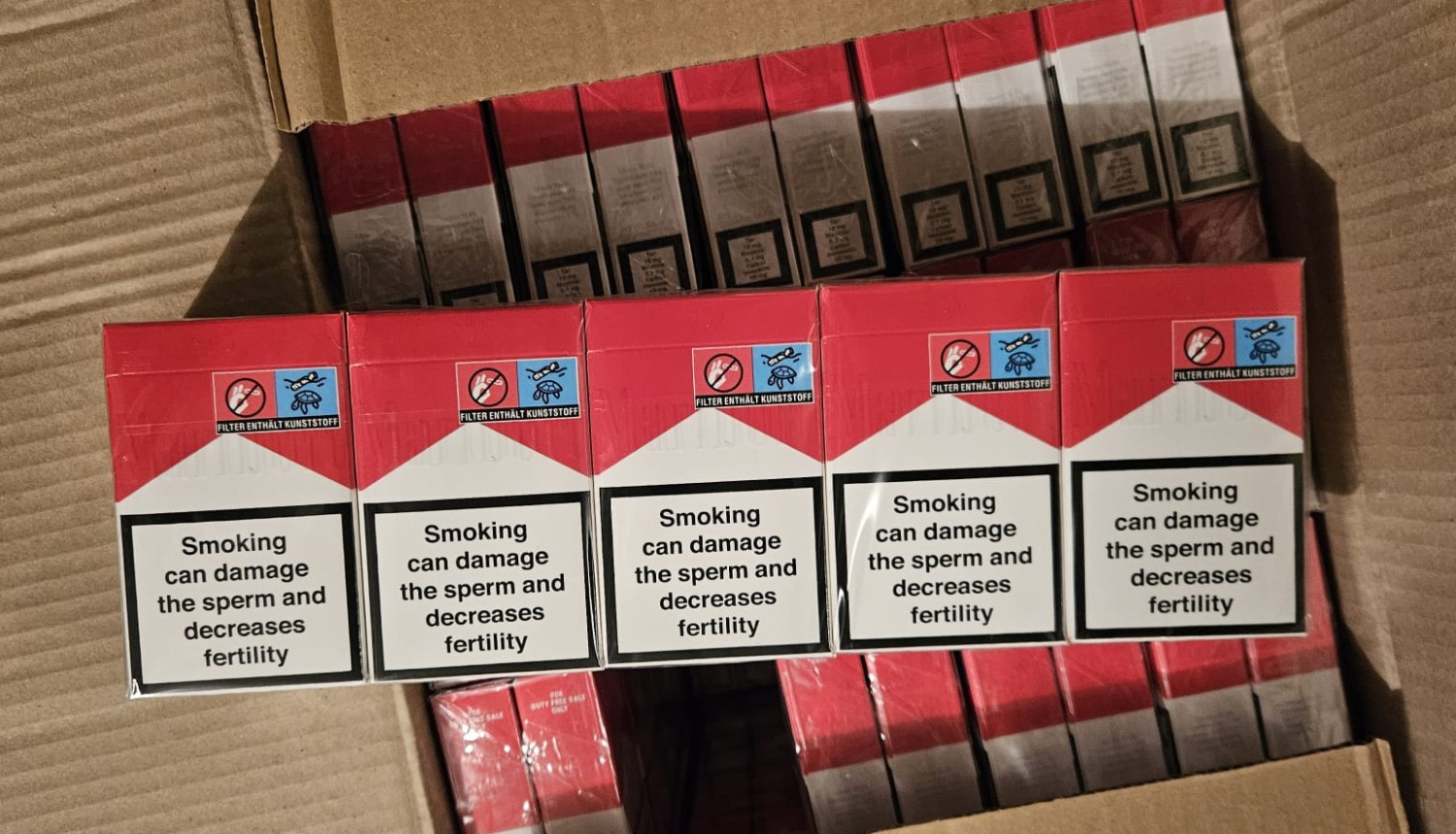 Uz kastes uzliktas piecas cigarešu paciņas sarkanā un baltā krāsā 