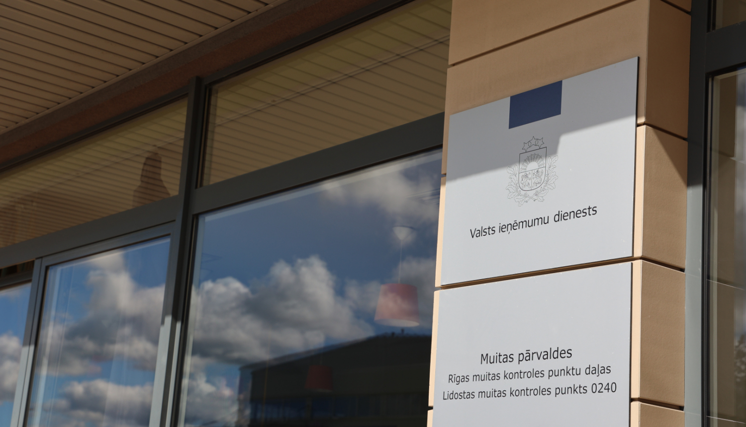 Pie ēkas fasādes piestiprinātas flīzes ar uzrakstiem, logos mākoņu atspīdums