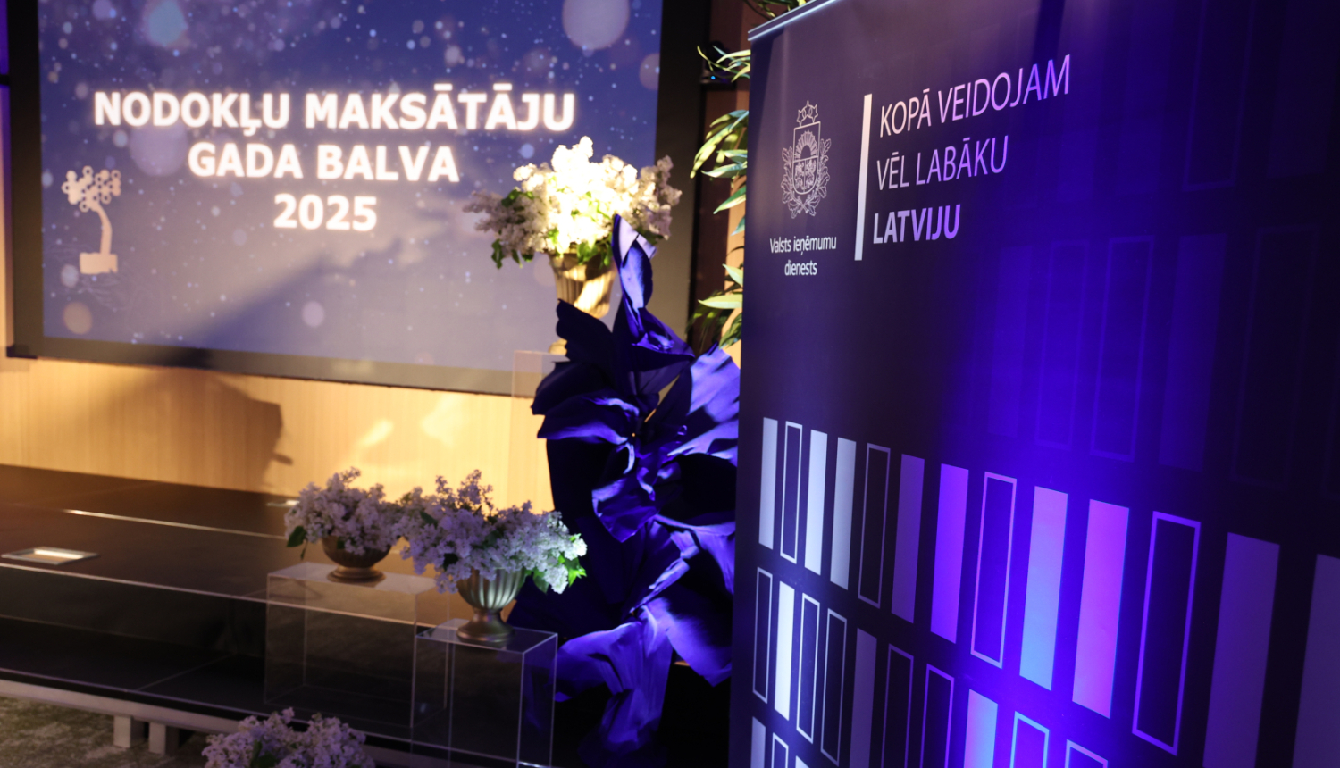 Banneris ar uzrakstu "Nodokļu maksāt'ju gada balva" ziedu dekorācijas