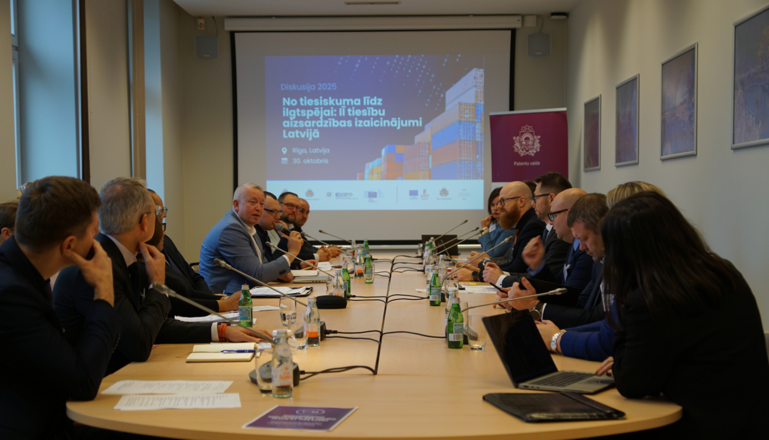 Abās galda pusēs sēž cilvēki, galā ekrāns un banneris