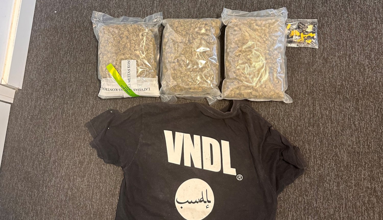 Uz grīdas trīs nelieli caurspīdīgi plēves iepakojumi ar brūnas krāsas narkotiskajām vielām 