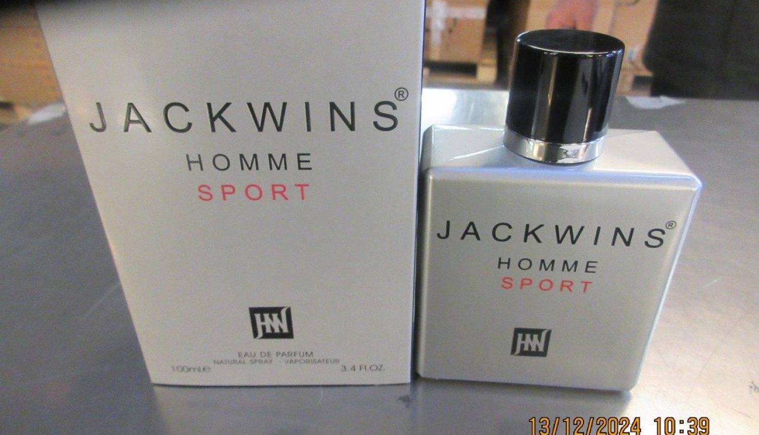 Uz galda pelēka smaržu pudelīte, blakus pelēkam smaržu iepakojumam ar melniem un sarkaniem uzrakstiem 