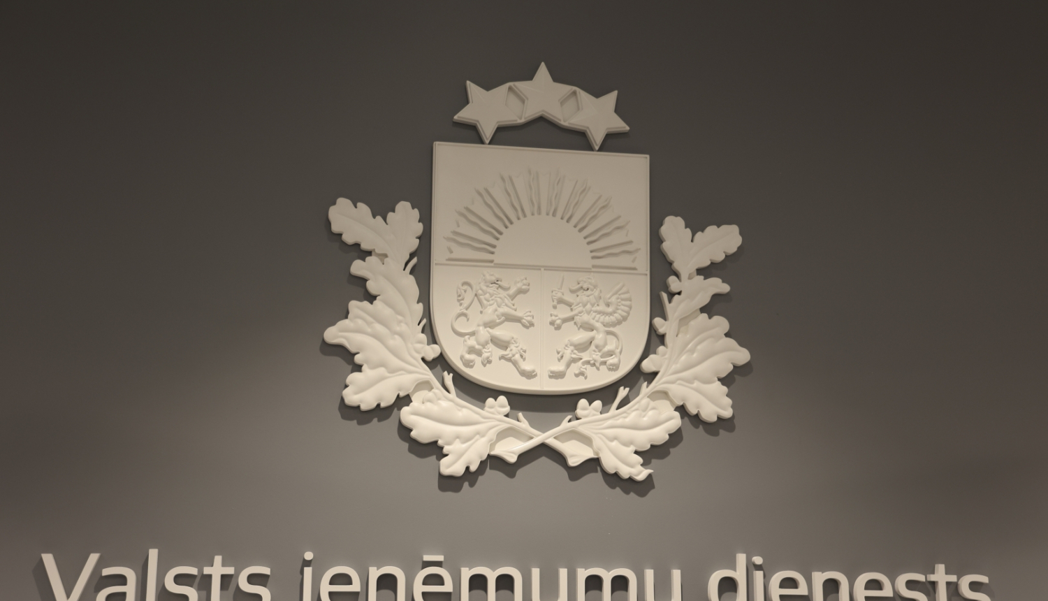 Balts ģērbonis un uzraksts Valsts ieņēmumu dienests uz pelēkas sienas