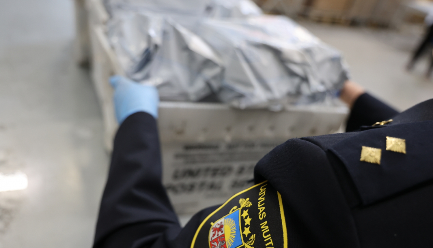 Priekšplānā muitas darbinieka žakete, rokās kaste ar pasta sūtījumiem