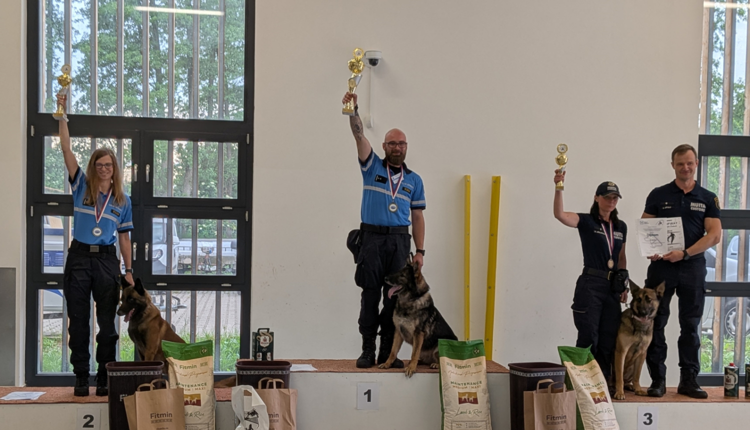 Uz pjedestāla stāv sacensību uzvarētāji – kinologi ar saviem darba suņiem