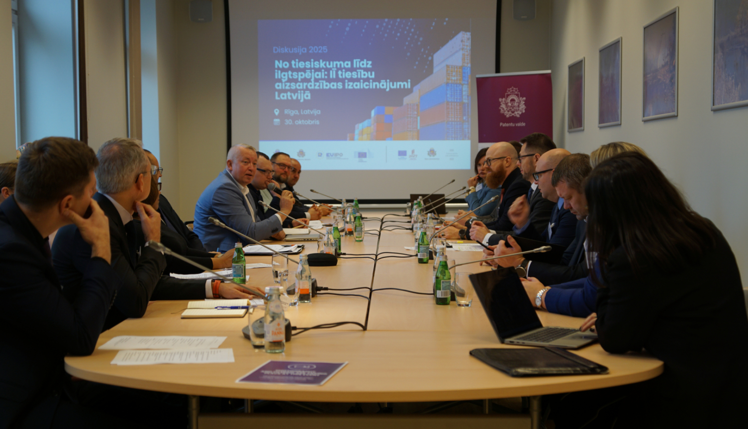 Abās galda pusēs sēž cilvēki, galā ekrāns un banneris